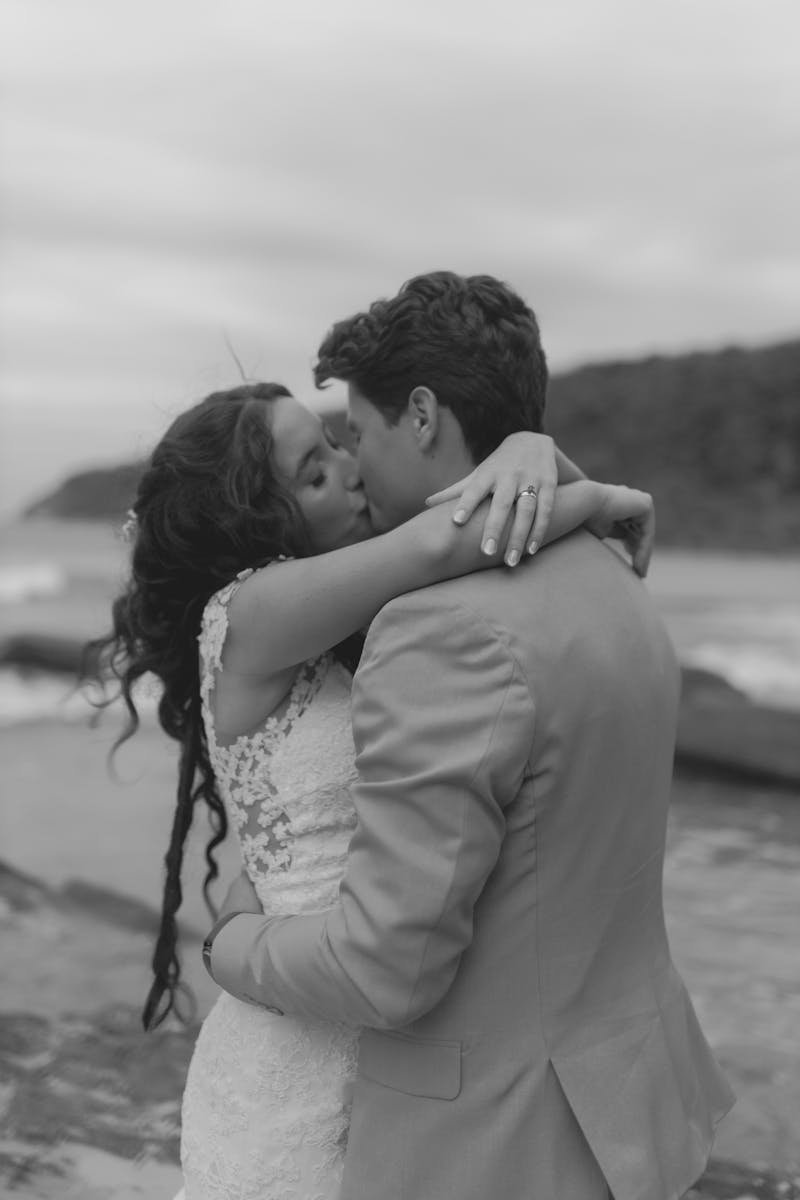 A couple in wedding attire shares a romantic kiss on a beach setting. Black and white photo.