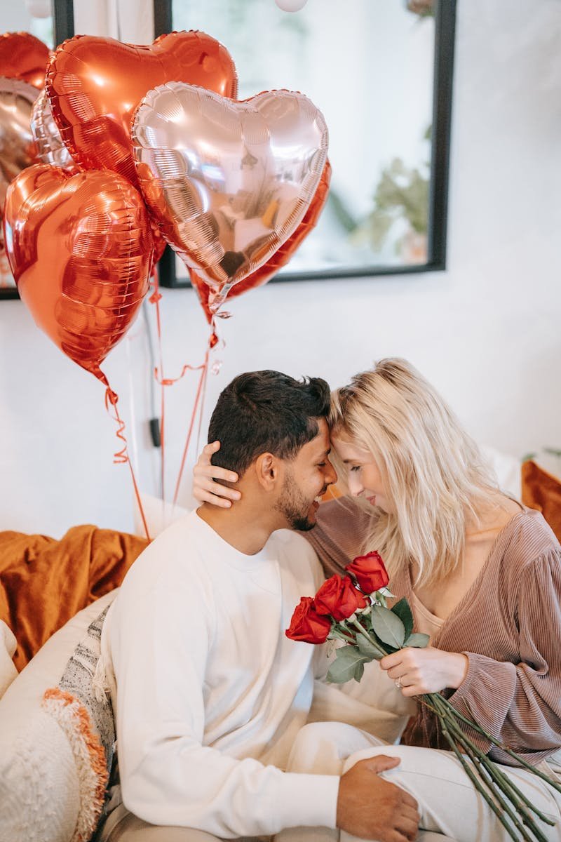 A romantic couple embraces indoors with heart-shaped balloons and roses, symbolizing love and affection.